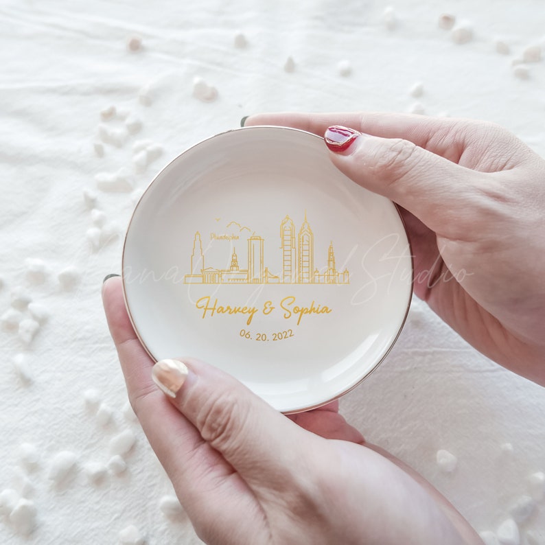 Op de afbeelding: Een witte keramische ringenschaal met een gouden rand, met een gouden skyline-ontwerp en de namen "Harvey & Sophia" met de datum "06.20.2022". De schaal wordt in twee handen gehouden.