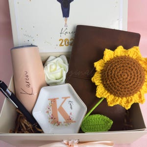 K&ouml;nnte beinhalten: Geschenkbox mit personalisiertem Becher, Stift, Notizbuch und Schmuckschale. Der Becher und die Schale sind mit dem Namen "Karen" beschriftet. Ein geh&auml;kelter Sonnenblume und eine wei&szlig;e Rose sind ebenfalls enthalten. Die Box hat ein Abschluss-Thema.