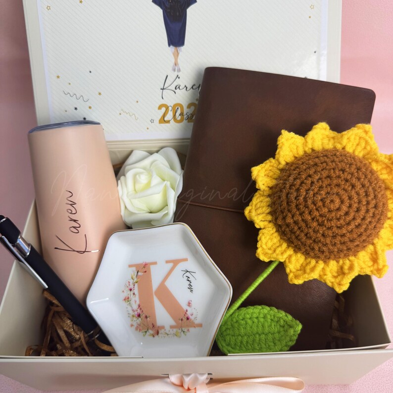 K&ouml;nnte beinhalten: Geschenkbox mit personalisiertem Becher, Stift, Notizbuch und Schmuckschale. Der Becher und die Schale sind mit dem Namen "Karen" beschriftet. Ein geh&auml;kelter Sonnenblume und eine wei&szlig;e Rose sind ebenfalls enthalten. Die Box hat ein Abschluss-Thema.