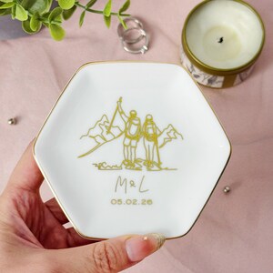 May include: White hexagonal ceramic trinket dish with a gold rim. The dish features a gold illustration of two hikers on a mountain, with the initials "M&L" and the date "05.02.26" below. A candle and rings are in the background.