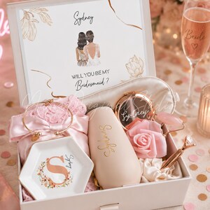 Op de afbeelding: Een witte geschenkdoos met een bruidsmeisje-aanzoekkaart met de naam Sydney, een roze satijnen badjas, een gouden armband, een zeshoekig keramisch schaaltje met een florale S, een beige beker, een compacte spiegel, een roos en een jade roller.