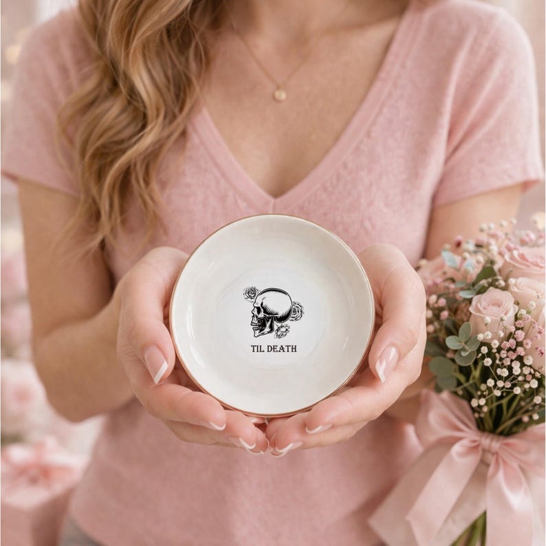 May include: A small, round, white ceramic trinket dish with a metallic gold rim. The center features a black line-art illustration of a human skull surrounded by roses, with the text "TIL DEATH" printed in black capital letters below it.