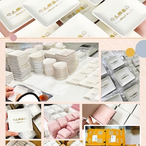 May include: A collection of white square ceramic dishes featuring a gold-colored graphic print of a house icon and the text "Always & Co". The dishes are shown in various stages of organization, including stacked piles, individual pieces, and packed boxes.