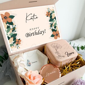 Puede incluir: Una caja de regalo rosa con un dise&ntilde;o floral y el texto "Katie &iexcl;Feliz cumplea&ntilde;os!" en la tapa. La caja contiene un plato de cer&aacute;mica blanco con el nombre "Laura" en &eacute;l, una caja de joyer&iacute;a rosa con el nombre "Laura" en ella, un espejo dorado con el nombre "Laura" en &eacute;l y una rosa rosa.
