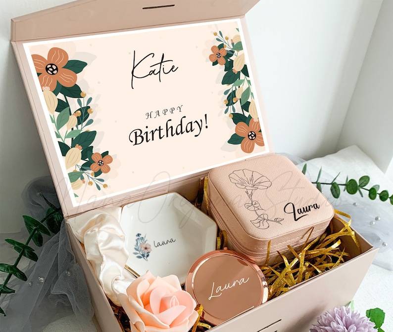 Puede incluir: Una caja de regalo rosa con un dise&ntilde;o floral y el texto "Katie &iexcl;Feliz cumplea&ntilde;os!" en la tapa. La caja contiene un plato de cer&aacute;mica blanco con el nombre "Laura" en &eacute;l, una caja de joyer&iacute;a rosa con el nombre "Laura" en ella, un espejo dorado con el nombre "Laura" en &eacute;l y una rosa rosa.