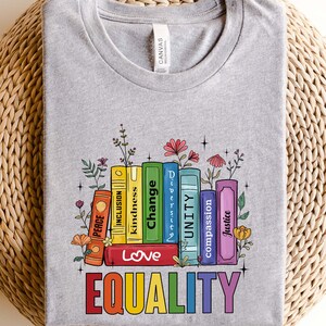 May include: A grey t-shirt with a stack of colorful books that have words like "Love", "Equality", "Change", "Justice", "Compassion", "Unity", "Diversity", "Kindness", "Inclusion", and "Peace" printed on the spines. The books are surrounded by flowers.