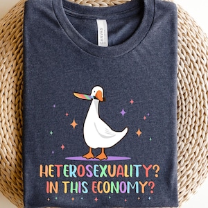 K&ouml;nnte beinhalten: Ein dunkelblaues T-Shirt mit einer wei&szlig;en Gans, die einen Regenbogen-Lolli im Schnabel h&auml;lt. Der Text "Heterosexuality? In this economy?" ist unter der Gans aufgedruckt.