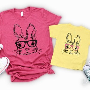 May include: Two t-shirts featuring a black and white illustration of a bunny wearing pink glasses. The larger shirt is pink and the smaller shirt is yellow. Both shirts are laid out on a white background with white sneakers.