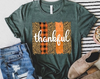 May include: A green t-shirt with a graphic design of orange, black, and white leopard print, buffalo plaid, and brushstrokes. The word "thankful" is written in white on the graphic.