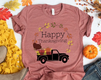 Puede incluir: Una camiseta color malva con un camión negro que lleva calabazas y un pavo. El camión está rodeado de una corona de hojas de otoño. El texto "Happy Thanksgiving" está impreso en la camiseta.