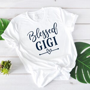 May include: White t-shirt with the text "Blessed Gigi" in navy blue lettering with a small heart graphic.