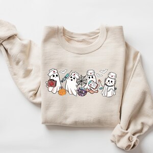 May include: A beige sweatshirt with a graphic of four ghosts dressed as nurses. The ghosts are holding medical supplies and a pumpkin. The text "Boo Crew" is printed on the sweatshirt.