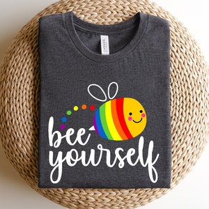 May include: A dark gray t-shirt with a white graphic of a rainbow bee and the text "bee yourself".