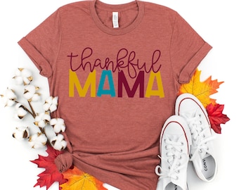 May include: A rust-colored t-shirt with the text "thankful MAMA" in colorful letters. The letters are yellow, blue, maroon, and yellow. The shirt is laid out on a white background with fall leaves and cotton bolls.