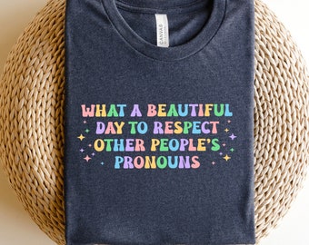 May include: A dark gray t-shirt with the text "WHAT A BEAUTIFUL DAY TO RESPECT OTHER PEOPLE'S PRONOUNS" in rainbow colors with glitter accents.