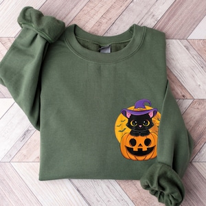 Puede incluir: Una sudadera verde con un gato negro con un sombrero de bruja dentro de una calabaza de Halloween.