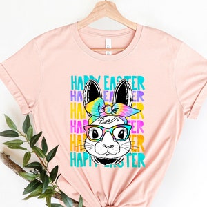 May include: A pink t-shirt with a tie-dye bunny wearing glasses and a headband. The text "Happy Easter" is repeated in rainbow colors.