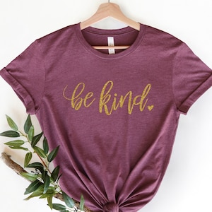 May include: A maroon t-shirt with the text "be kind" in gold glitter lettering.
