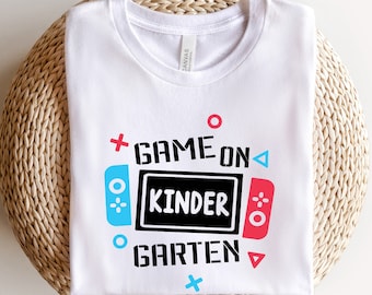 Puede incluir: Camiseta blanca con un diseño gráfico de un mando de videojuego con el texto "Game On Kinder Garden".