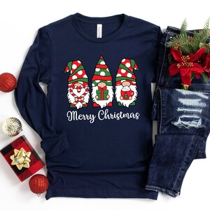May include: A navy blue long-sleeved shirt with a Christmas gnome design. The gnomes are wearing red and white hats, and holding a candy cane, a gift box, and a cup of hot chocolate. The text "Merry Christmas" is printed below the gnomes.