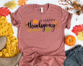 Könnte beinhalten: Ein mauvefarbenes T-Shirt mit dem Text "Happy Thanksgiving" in schwarzer Schrift mit einem Kürbis und Herbstblättern. Das Shirt liegt auf einer weißen Oberfläche mit einem gelben Strickpullover, braunen Stiefeln, einer blauen Jeans und Herbstblättern.
