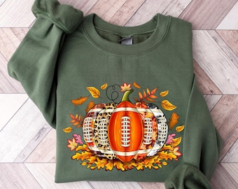 May include: A green sweatshirt with a graphic of a pumpkin made of footballs surrounded by fall leaves.