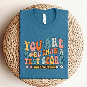 May include: A blue t-shirt with a retro-style graphic that says "You are more than a test score" with a yellow pencil and a flower. The hashtag #Testday is below the pencil.
