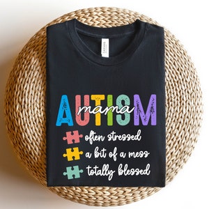 K&ouml;nnte beinhalten: Schwarzes T-Shirt mit dem Text "AUTISM mama" in Regenbogenfarben. Unter dem Text befinden sich drei Puzzle-Teile mit dem Text "oft gestresst", "ein bisschen ein Chaos" und "total gesegnet".