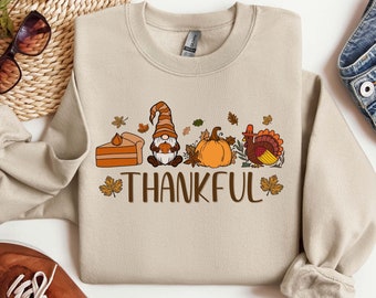May include: A beige sweatshirt with a Thanksgiving-themed design. The design features a gnome holding a pie, a pumpkin, and a turkey. The text "THANKFUL" is printed below the design.