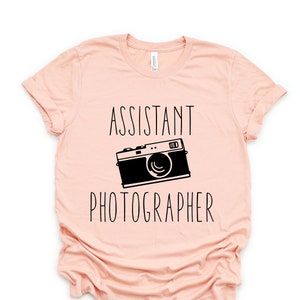 May include: A light pink t-shirt with black text that reads "Assistant Photographer" and a black illustration of a vintage camera.