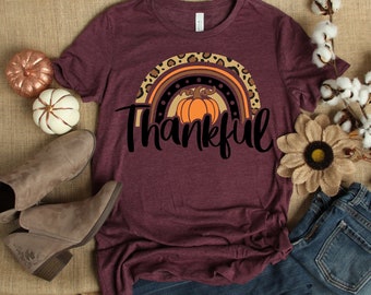 May include: A maroon t-shirt with a rainbow design featuring a pumpkin and the word "Thankful". The rainbow is decorated with leopard print and black dots.