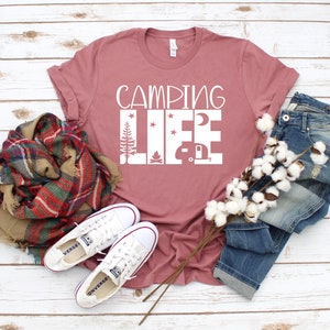 May include: A dusty rose t-shirt with the text "Camping Life" in white. The text is surrounded by images of a campfire, a camper, a moon, and stars. The t-shirt is laid out on a white wooden surface with a pair of white sneakers, a plaid scarf, and a pair of blue jeans.