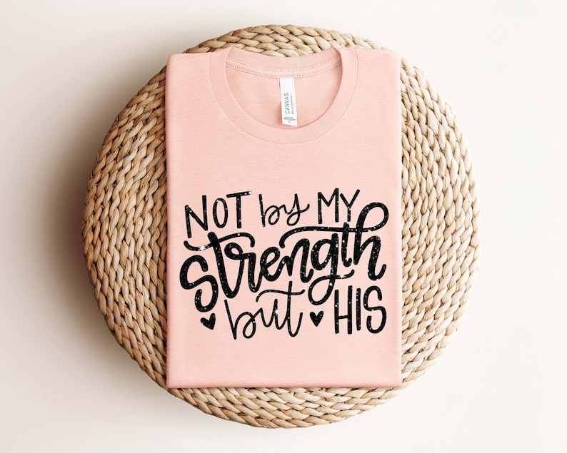 Op de afbeelding: Een roze T-shirt met de tekst "NOT by MY Strength but HIS" in zwarte letters met een glittereffect.