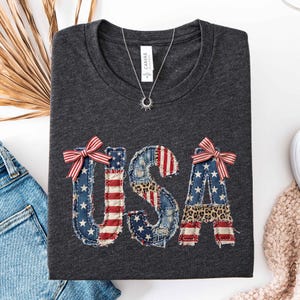 May include: A dark charcoal gray t-shirt featuring the letters USA in a distressed, patchwork-style print. The letters incorporate American flag patterns, denim textures, and leopard print accents, topped with two red and white striped bows.
