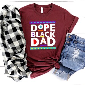 May include: A maroon t-shirt with the text "Dope Black Dad" in colorful block letters. The shirt is laid out on a white wooden surface with a black and white plaid shirt and blue jeans.