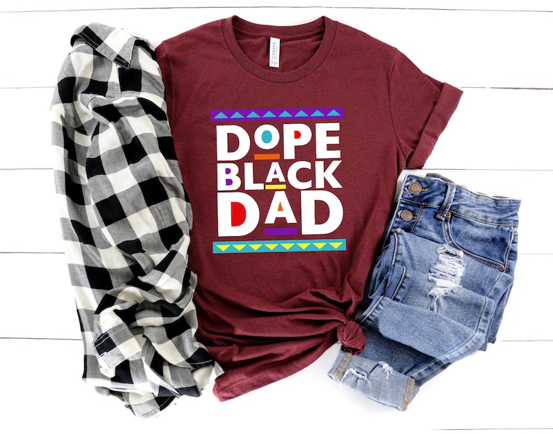 May include: A maroon t-shirt with the text "Dope Black Dad" in colorful block letters. The shirt is laid out on a white wooden surface with a black and white plaid shirt and blue jeans.