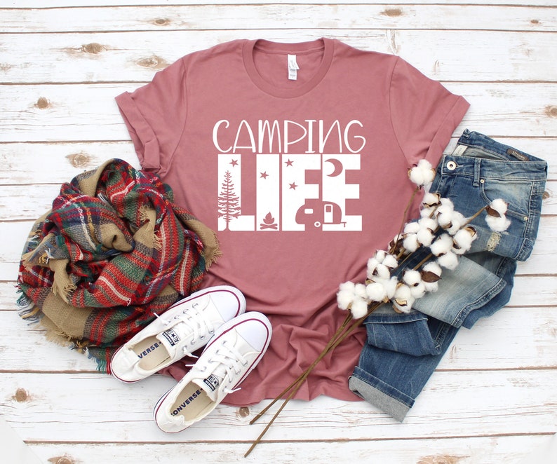 May include: A dusty rose t-shirt with the text "Camping Life" in white. The text is surrounded by images of a campfire, a camper, a moon, and stars. The t-shirt is laid out on a white wooden surface with a pair of white sneakers, a plaid scarf, and a pair of blue jeans.