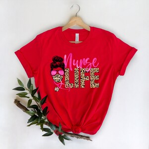 May include: Red t-shirt with a pink and black leopard print design that says "Nurse Life". The design features a woman with a bun in her hair wearing a face mask and sunglasses.