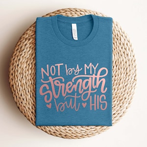 Op de afbeelding: Een blauw t-shirt met de tekst "NOT by MY strength but HIS" in roze letters.