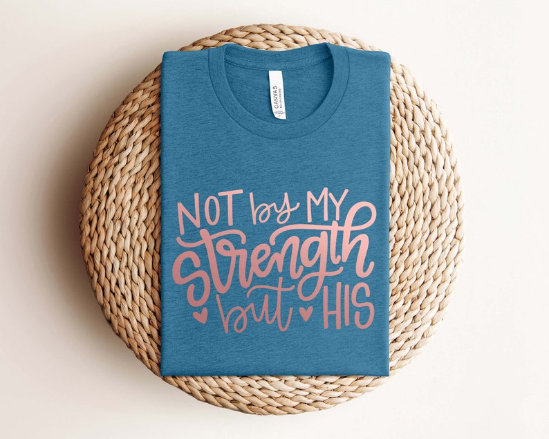 Op de afbeelding: Een blauw t-shirt met de tekst "NOT by MY strength but HIS" in roze letters.