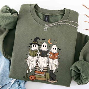 May include: An olive green crewneck sweatshirt featuring a graphic print of three ghosts wearing hats and reading books while sitting on a stack of books. The design includes a black cat, a pumpkin, a candle, bats, and a crescent moon in the background.