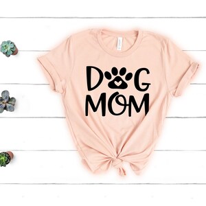 Pode incluir: Uma camiseta rosa claro com um gr&aacute;fico preto que diz "Dog Mom" com uma pegada de pata e um cora&ccedil;&atilde;o no meio da palavra "Dog".