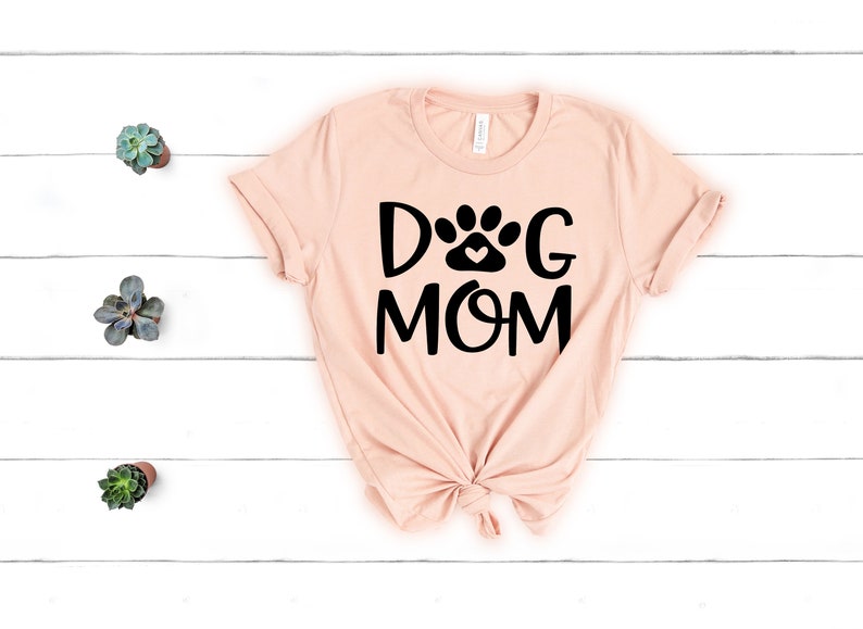 Pode incluir: Uma camiseta rosa claro com um gr&aacute;fico preto que diz "Dog Mom" com uma pegada de pata e um cora&ccedil;&atilde;o no meio da palavra "Dog".