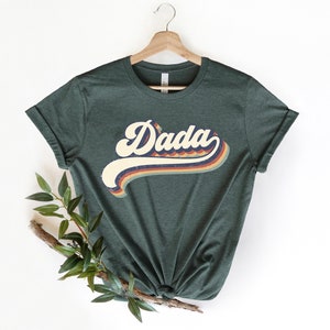 Może przedstawiać: Zielony T-shirt z grafiką w stylu retro, na kt&oacute;rej widnieje napis "Dada" w kolorowym, falistym foncie.