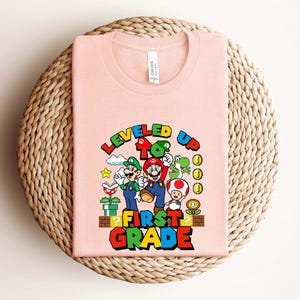Puede incluir: Una camiseta rosa claro con un gr&aacute;fico colorido de Mario, Luigi y otros personajes de la serie de videojuegos Super Mario. El texto "LEVELED UP TO FIRST GRADE" se muestra en una fuente vibrante y multicolor.