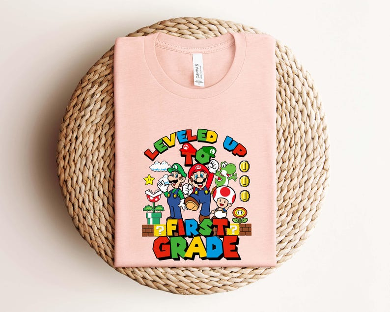 Puede incluir: Una camiseta rosa claro con un gr&aacute;fico colorido de Mario, Luigi y otros personajes de la serie de videojuegos Super Mario. El texto "LEVELED UP TO FIRST GRADE" se muestra en una fuente vibrante y multicolor.