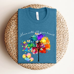 May include: A teal blue t-shirt with a colorful watercolor tree and butterflies. The text "Love with all your heart" is written in white script on the shirt.