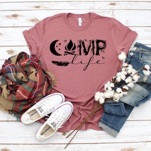 May include: A mauve t-shirt with a black graphic that says "Camp Life" with a campfire, moon, and feathers. The shirt is laid out on a white wood background with a plaid scarf, white cotton stems, and a pair of white canvas shoes.