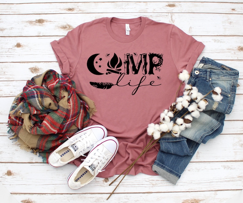 May include: A mauve t-shirt with a black graphic that says "Camp Life" with a campfire, moon, and feathers. The shirt is laid out on a white wood background with a plaid scarf, white cotton stems, and a pair of white canvas shoes.