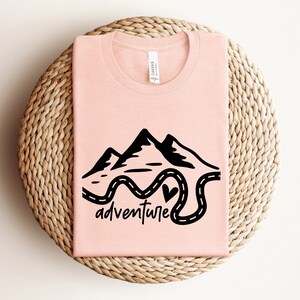 以下が含まれることがあります： 山と曲がりくねった道の黒いグラフィックが描かれたピンクのTシャツ。道の終わりにはハートマークがあります。グラフィックの下には「adventure」という文字があります。